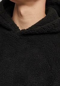 Hoodie en polaire noire avec une surface douce et texturée, présentant une capuche et des détails côtelés le long de l'encolure.