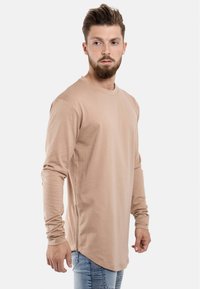 Cămașă bej cu mâneci lungi, realizată dintr-un material moale și neted, având un guler rotund și detalii cu slituri laterale pentru un design modern. Asortată cu blugi albaștri deschis.
