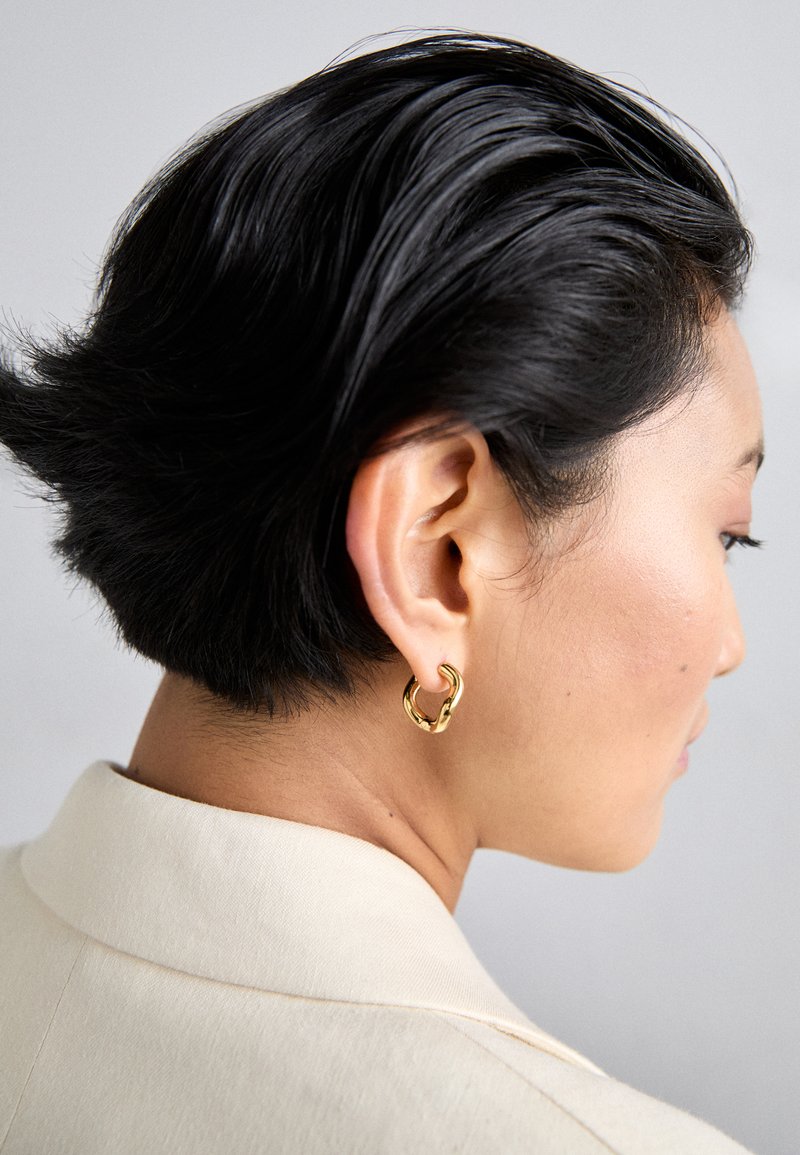 Goldene Metall-Hoop-Ohrringe, getragen in kurzen schwarzen Haaren, seitlich dargestellt, mit glatter, polierter Textur und modernem Design.