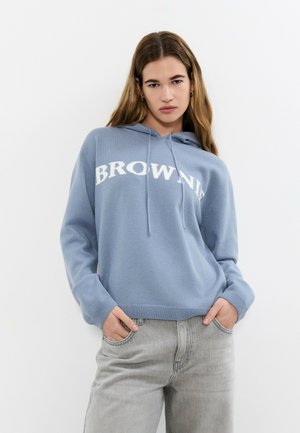 Joven con sudadera con capucha azul con el texto "BROWNIE", posando con las manos en los bolsillos de sus vaqueros gris claro frente a un fondo blanco.