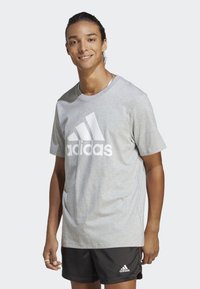 Šedé tričko s krátkým rukávem a velkým bílým logem Adidas na přední straně, vyrobené z měkké tkaniny. Kombinováno s černými šortkami, které mají malé logo.
