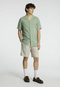 Camicia verde chiaro a maniche corte con bottoni, abbinata a pantaloni corti beige. Modello con calze bianche e scarpe marroni scure.