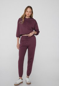 Burgundowy skrócony hoodie z kapturem na sznurku, w połączeniu z dopasowanymi dresami ze zwężającymi się nogawkami i elastycznym pasem, noszonymi z białymi trampkami.