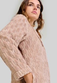 Mujer vistiendo una chaqueta de felpa texturizada en color beige con mangas largas, de pie frente a un fondo gris claro liso.