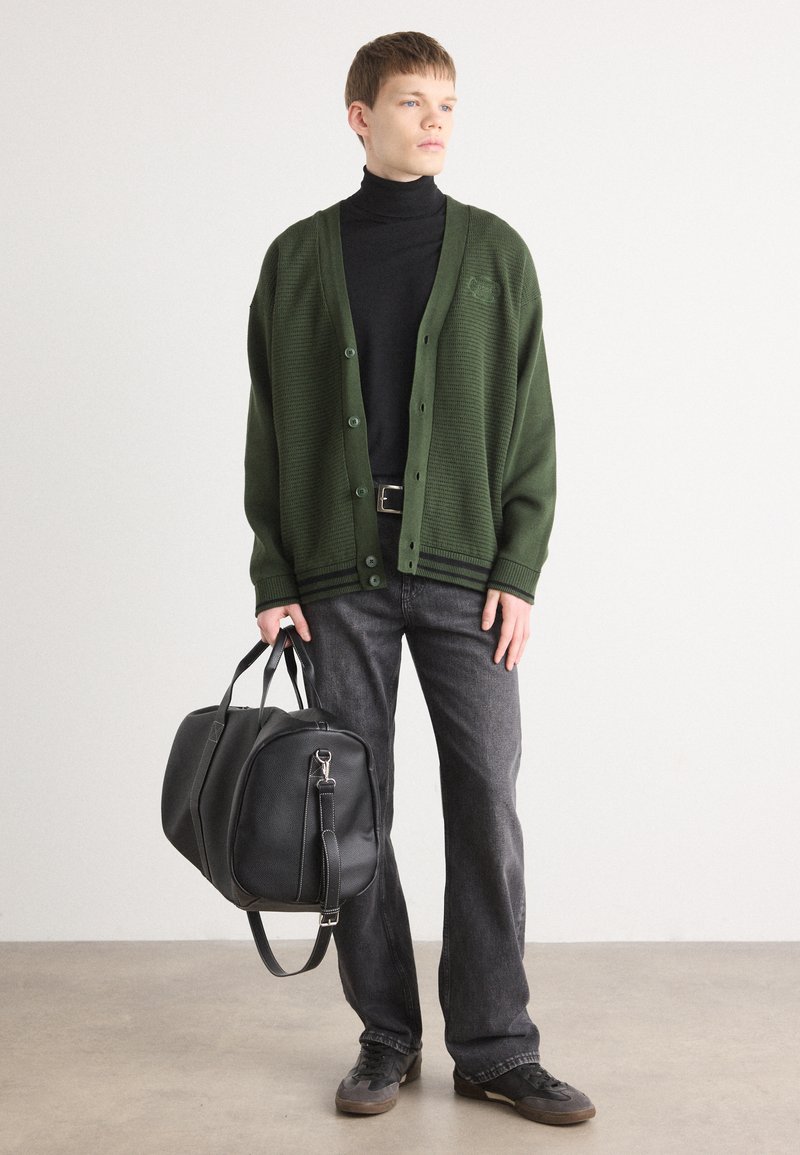 Zielony sweter z dzianiny z zapięciem na guziki, czarny golf, szare dżinsy oraz czarna skórzana torba z podwójnymi uchwytami i paskiem. Teksturowany materiał.