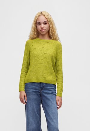 Jeune femme aux longs cheveux blonds bouclés portant un pull en maille vert citron et un jean bleu, debout devant un fond clair uni.