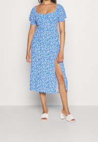 Blaues blumiges Midikleid mit Puffärmeln, quadratischem Ausschnitt und seitlichem Schlitz. Das Modell trägt weiße Blockabsatzsandalen. Weicher Stoff mit kleinen Gänseblümchen.