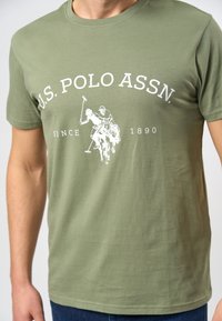 Olīvzaļš kokvilnas T-krekls ar baltu grafiku, kurā attēloti divi polo spēlētāji jāšanā, ar tekstu "U.S. POLO ASSN." un "SINCE 1890".