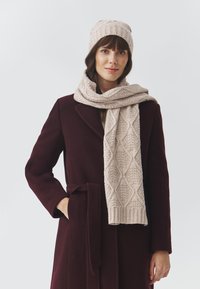 Cappello a maglia beige e sciarpa con motivi a diamante testurizzati, abbinati a un cappotto bordeaux scuro. La sciarpa è avvolta e drappeggiata in modo morbido.