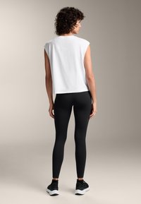 Weißes, ärmelloses Oberteil kombiniert mit schwarzen Leggings, mit lässiger Passform und glatter Textur. Model trägt schwarze Sneaker, von hinten gesehen.
