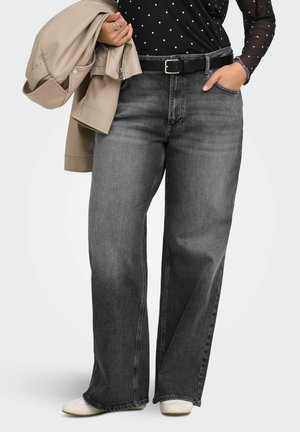 Personne portant un jean gris taille haute à jambes larges, ceinture noire, chaussures blanches, tenant une veste beige sur un bras, une main dans la poche, portant un haut noir à pois.