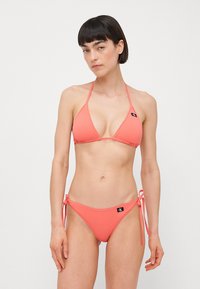 Korálový bikini z mäkkého materiálu; obsahuje trojuholníkový top s tenkými ramienkami a nízko strihané nohavičky so šnúrovaním po bokoch a logom.