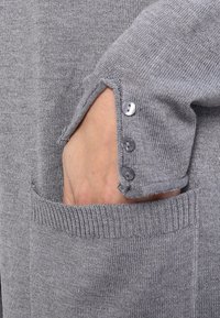 Maglione grigio a maglia con polsini a coste e tasca frontale. Caratterizzato da tre bottoni grigi sul polsino per una vestibilità regolabile. Texture morbida.