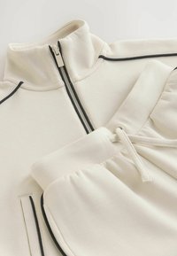 Ljusbeige zip-up sweatshirt med hög krage och svarta detaljer; matchas med byxor i samma färg med elastisk midja och dragsko.