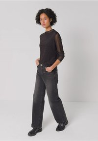 Czarna, przezroczysta bluzka z długim rękawem w połączeniu z ciemnoszarymi, szerokimi spodniami o wysokim stanie. Modelka stoi z rękami w kieszeniach, w czarnych butach.