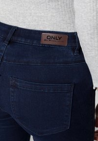 Dark blue denim jeans with a high-waisted design. Features a stitched back pocket and a brown leather-like tag with "ONLY" branding.
