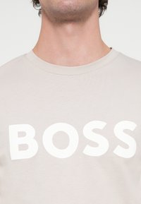 Ανοιχτό μπεζ βαμβακερό t-shirt με έντονο, κεφαλαίο λευκό κείμενο "BOSS" στο στήθος. Λαιμόκοψη σε στρογγυλό σχήμα με κοντά μανίκια.