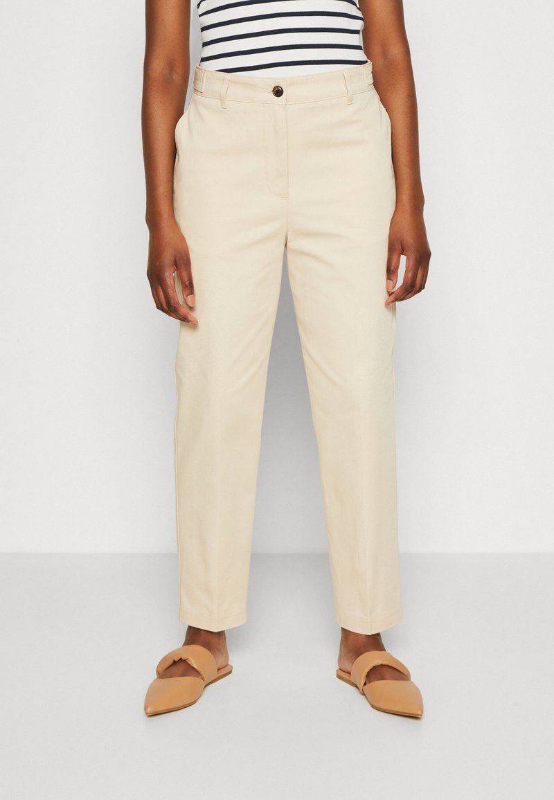 Personne portant un pantalon droit taille haute beige, des chaussures à enfiler à bout pointu couleur sable et un haut sans manches rayé noir et blanc.