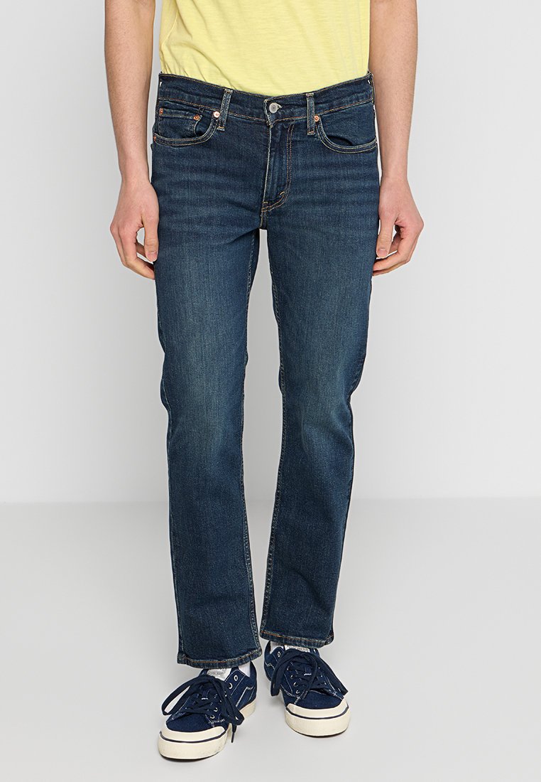 Dunkelblaue Denim-Jeans mit geradem Bein, klassischem Fünf-Taschen-Design und Knopfverschluss. Kombiniert mit navyfarbenen Turnschuhen und einem gelben Hemd.