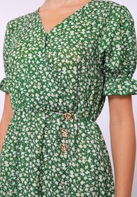 Grünes Blumenkleid mit V-Ausschnitt, kurzen Puffärmeln, tailliertem Schnitt und drei goldenen Knopfakzenten. Leichtes Material mit einem dezenten Glanz.