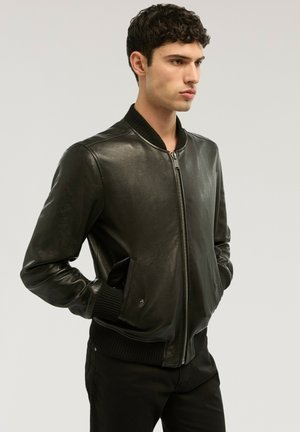 Giovane uomo con capelli ricci che indossa una giacca bomber di pelle nera con polsini e collo a costine, mani in tasca, rivolto verso destra.