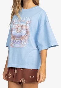 Camiseta de corte oversized color azul claro con un gráfico frontal que presenta un sol, flores y olas estilizadas en suaves tonos pastel. Mangas cortas y ajuste relajado.