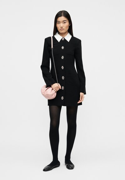 Γυναίκα που φοράει μαύρο φόρεμα με λευκό γιακά, διακοσμητικά κουμπιά, μαύρο κολάν, μαύρες φλατ παπούτσια και κρατά μια μικρή ροζ τσάντα ώμου.