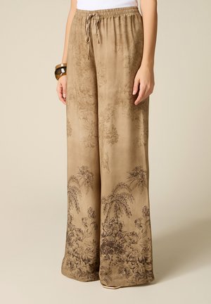 Pantaloni beige larghi con motivi scuri floreali e di palme, vita elastica con coulisse, indossati con una maglia bianca e braccialetti marroni.