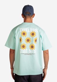 Mahan vihreä puuvillapaita, jossa on auringonkukka grafiikkakuosi selkäpuolella, tekstillä "KOHTI AURINKOA" ylhäällä ja siihen liittyvillä lauseilla alapuolella.