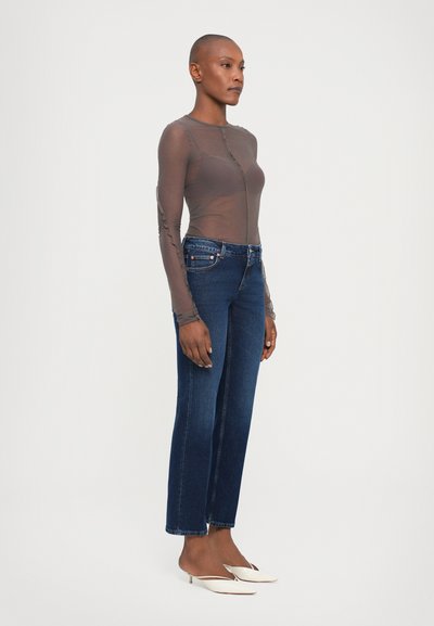 Long-sleeve, sheer brown top with a round neckline, paired with dark blue straight-leg jeans and white open-toe heels. Simple design.