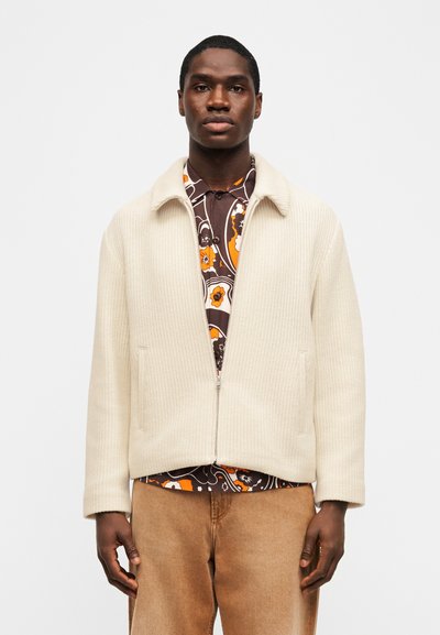 Άνδρας που φοράει εκρού πλεκτό μπουφάν με φερμουάρ πάνω από πουκάμισο με καφέ, πορτοκαλί και λευκό φλοράλ σχέδιο, και ανοιχτό καφέ παντελόνι, στέκεται μπροστά σε λευκό φόντο.