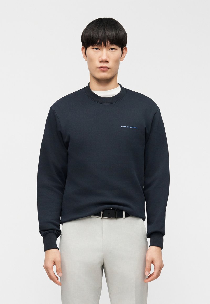 Μπλε σκούρο φούτερ με στρογγυλή λα ιμόκοψη και μακριά μανίκια. Διαθέτει απλό λογότυπο κειμένου στο αριστερό στήθος. Συνδυάζεται με ανοιχτό γκρι παντελόνι.