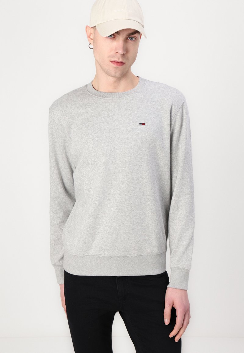 Szary sweter z długimi rękawami i ściągaczem, z małym czerwono-biało-niebieskim logo na klatce piersiowej. Uzupełniony czarnymi spodniami i jasną czapką.