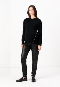Svart strikket genser med sidedetaljer, kombinert med svarte faux skinnbukser og skinnende svarte sko. Modellen står på en hvit bakgrunn.