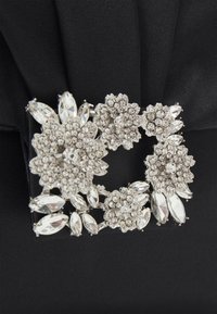 Srebrna broszka w kształcie kwiatów z różnorodnie ukształtowanymi kryształami na czarnym tle z tkaniny. Posiada skomplikowane detale i błyszczące wykończenie.