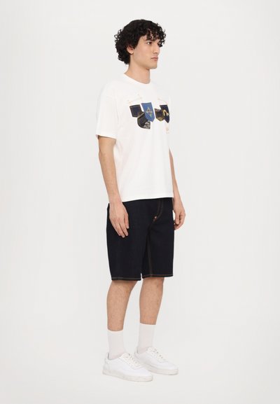 T-shirt blanche avec des éléments graphiques bleus et jaunes, associée à un short en denim foncé et des baskets blanches. Cheveux bouclés visibles.