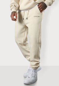 Beige Sweatpants mit relaxed Fit, ausgestattet mit einem Kordelzugbund, Seitentaschen und einem Logo-Detail am oberen linken Bein. Kombiniert mit weißen Sneakers.