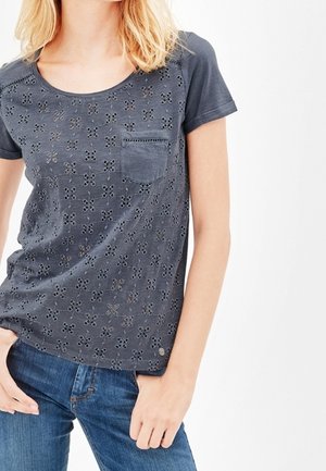 Kvinde iført en mørkegrå kortærmet skjorte med blomstret udskåret mønster og brystlomme, kombineret med blå jeans.