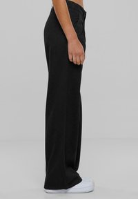 Persona con pantalones negros de cintura alta y pernera ancha y zapatillas blancas, de pie de lado contra un fondo gris liso.