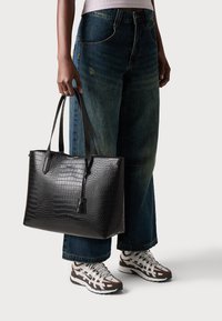 Sac fourre-tout noir texturé crocodile avec deux poignées, finition lisse, forme rectangulaire et petit accent pendu. Porté avec un jean à jambes larges et des baskets.