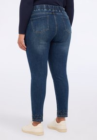 Jeans skinny in denim con una cintura elasticizzata, colore blu scuro, due tasche posteriori e decorazioni con borchie all'orlo della caviglia. Abbinati a scarpe bianche.