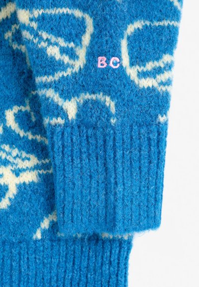 Manche de pull bleu tricoté avec motif abstrait blanc et lettres brodées roses "BC" au poignet.