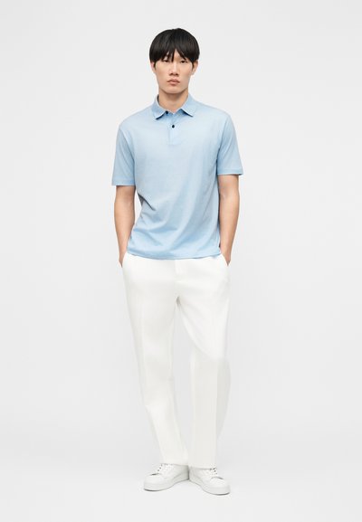 Νεαρός άνδρας στέκεται με τα χέρια στις τσέπες, φορώντας ένα γαλάζιο πόλο πουκάμισο, λευκό παντελόνι και λευκά αθλητικά παπούτσια, μπροστά σε απλό φόντο.