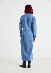 Denimkjole med lange ermer, krage og belte i midjen. Har en midtlengde og synlig søm, kombinert med knelangne svarte støvler.
