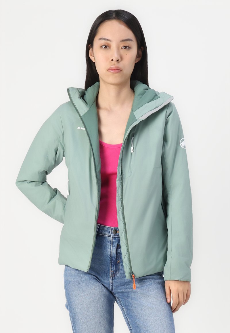 Jasnozielona kurtka ocieplana z kapturem, z zamkniętą kieszenią i kontrastującym szarym wnętrzem. Nosi ją osoba w różowej bluzce i dżinsowych spodniach.