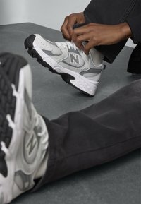 Baskets New Balance avec une tige en maille blanche et grise, semelle en caoutchouc noire, et le logo "N" emblématique. La personne ajuste les lacets.