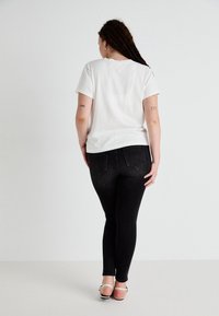 Frau mit geflochtenem Haar, die ein weißes T-Shirt, schwarze Skinny-Jeans und weiße Sandalen trägt, steht mit dem Rücken zur Kamera vor einem schlichten Hintergrund.