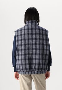 Tumši zils un pelēks pārbaudīts vestes modelis ar augstu apkakli, izgatavots no teksturēta auduma, ar elastīgu malu un rokas caurumiem, valkāta virs tumši zila garām piedurknēm krekla.