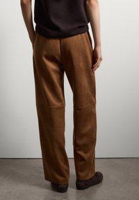 Pantalons larges en daim marron avec une texture lisse. Présente des coutures de poches et une coupe décontractée. Associés à des chaussures sombres, vus de l'arrière.