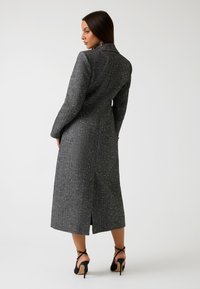 Cappotto lungo grigio con tessitura a trama e macchie bianche, dotato di colletto a punte e uno spacco posteriore per facilitare i movimenti.